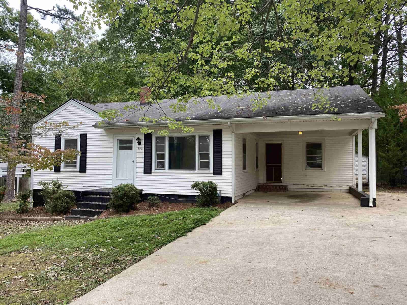 A single-story white house with black shutters and a covered porch surrounded by trees and a driveway on the left side.