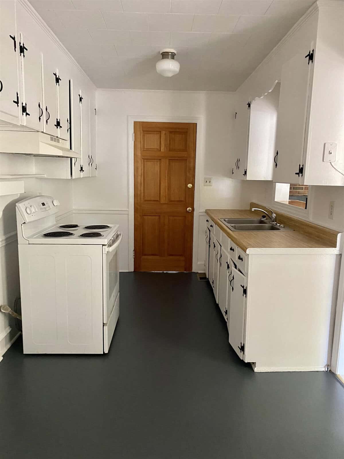 A small, traditional kitchen with white appliances, black and white cabinet hardware, a wooden countertop, and a wooden door at the end.