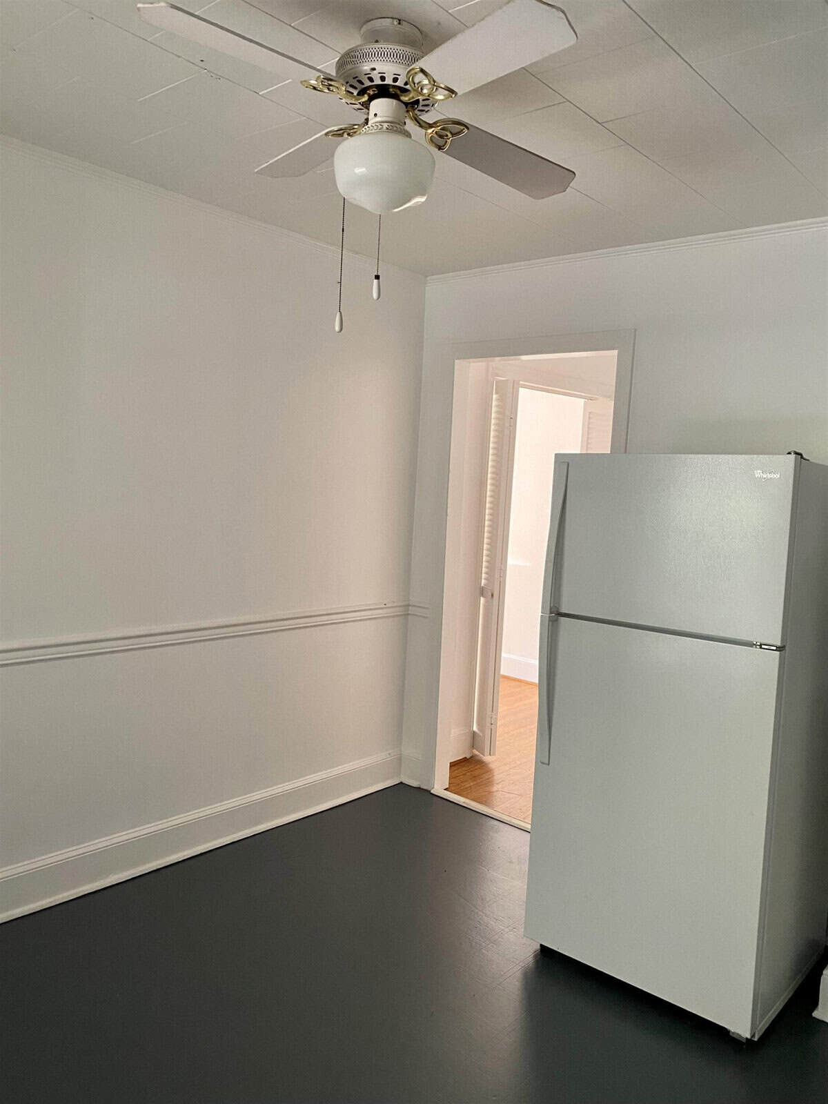 A sparsely furnished room with gray flooring, a single white refrigerator, and a ceiling fan.