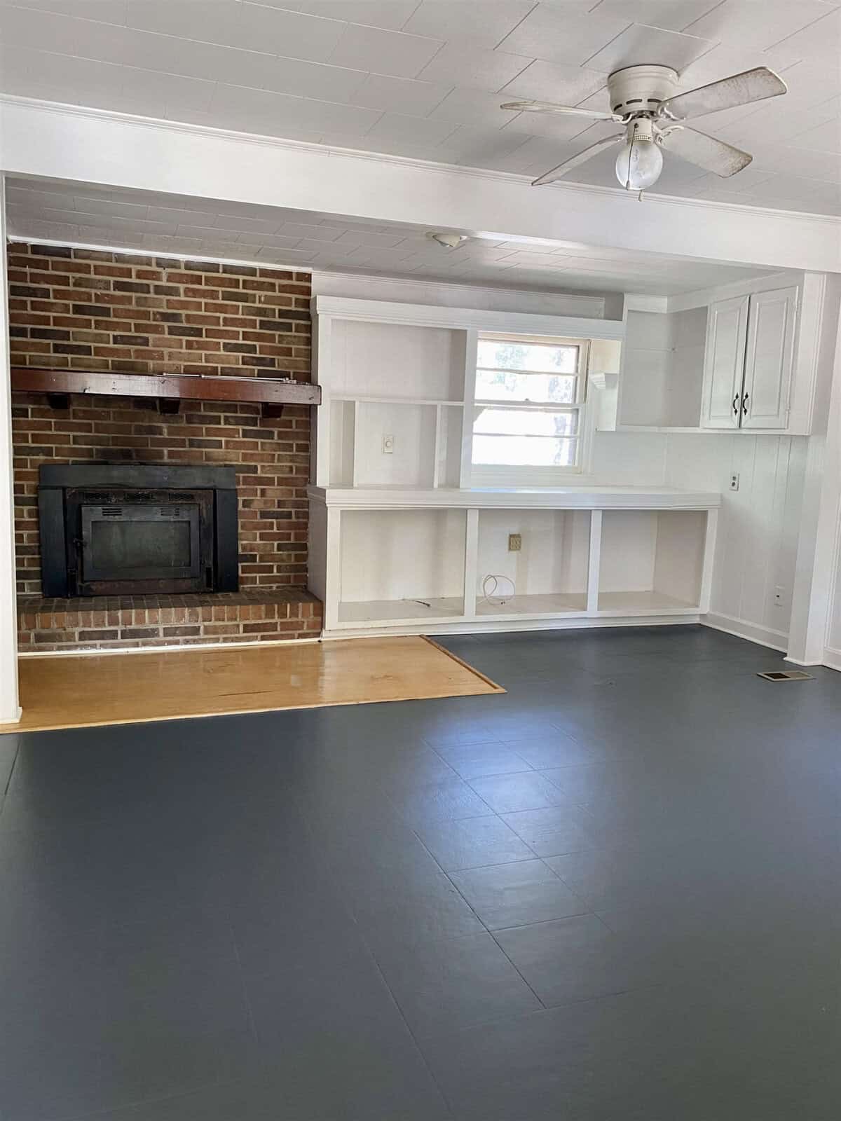An empty room featuring a brick fireplace centered on one wall, flanked by white built-in shelves and cabinets, with a gray floor and a ceiling fan above.