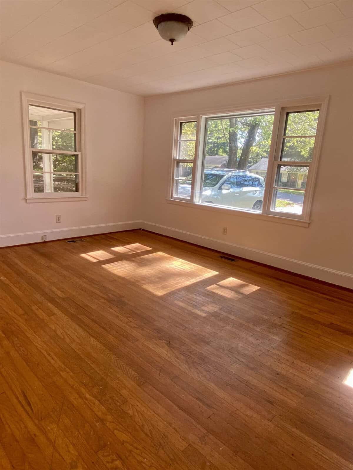 An empty room with hardwood floors, one small and one large window letting in natural light, and a simple light fixture on the ceiling.
