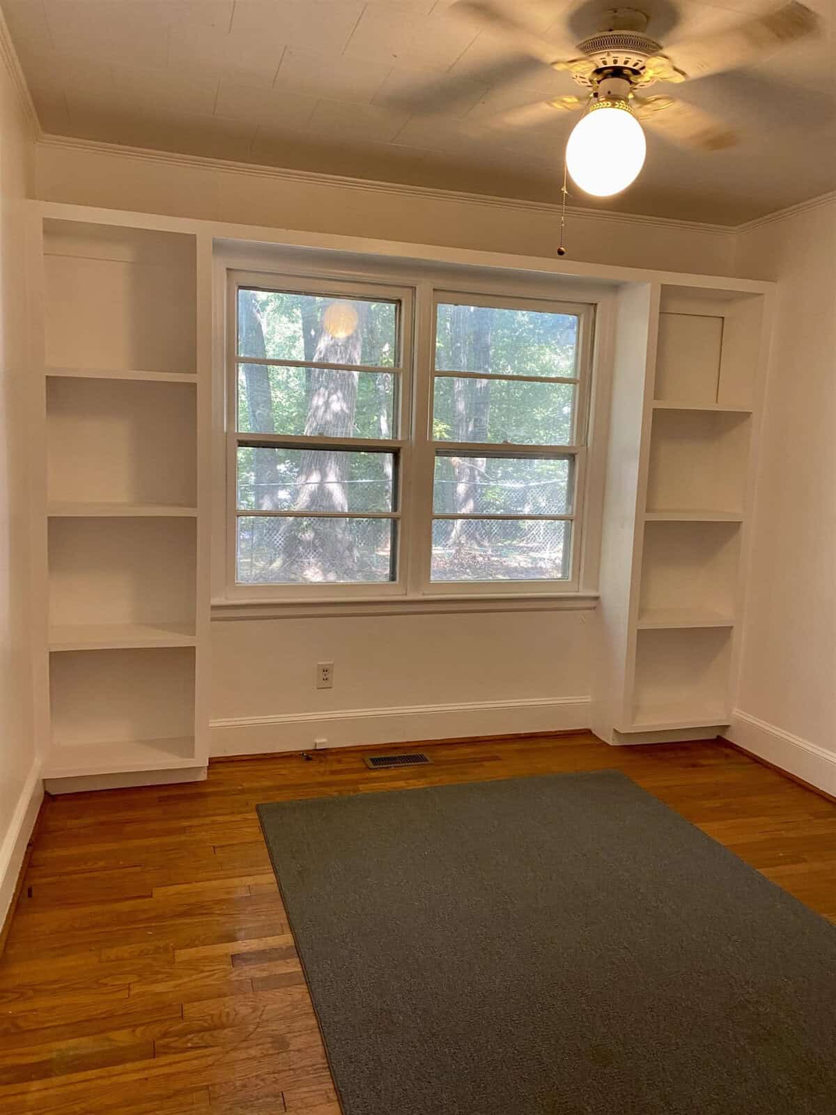 An empty room with hardwood floors, a gray area rug, white built-in bookshelves flanking a window, and a ceiling light fixture.