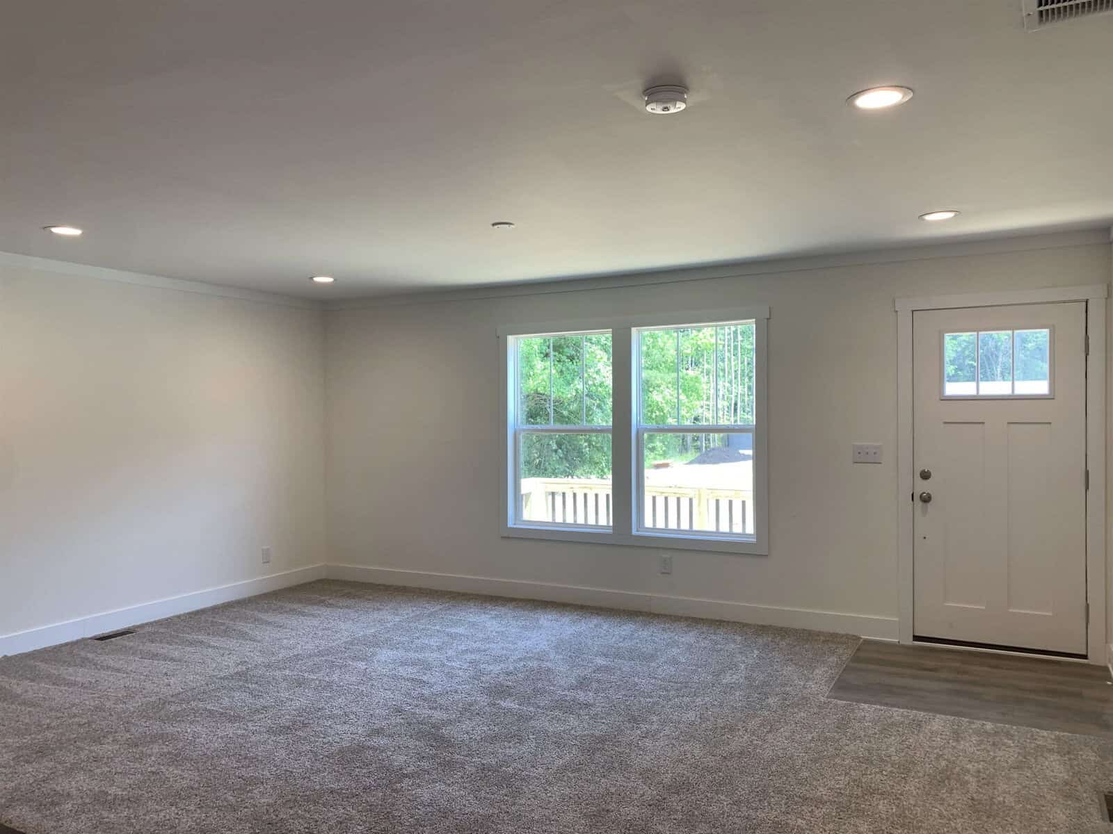 An empty room with gray carpeting, white walls with one window, and a front door with a glass panel.