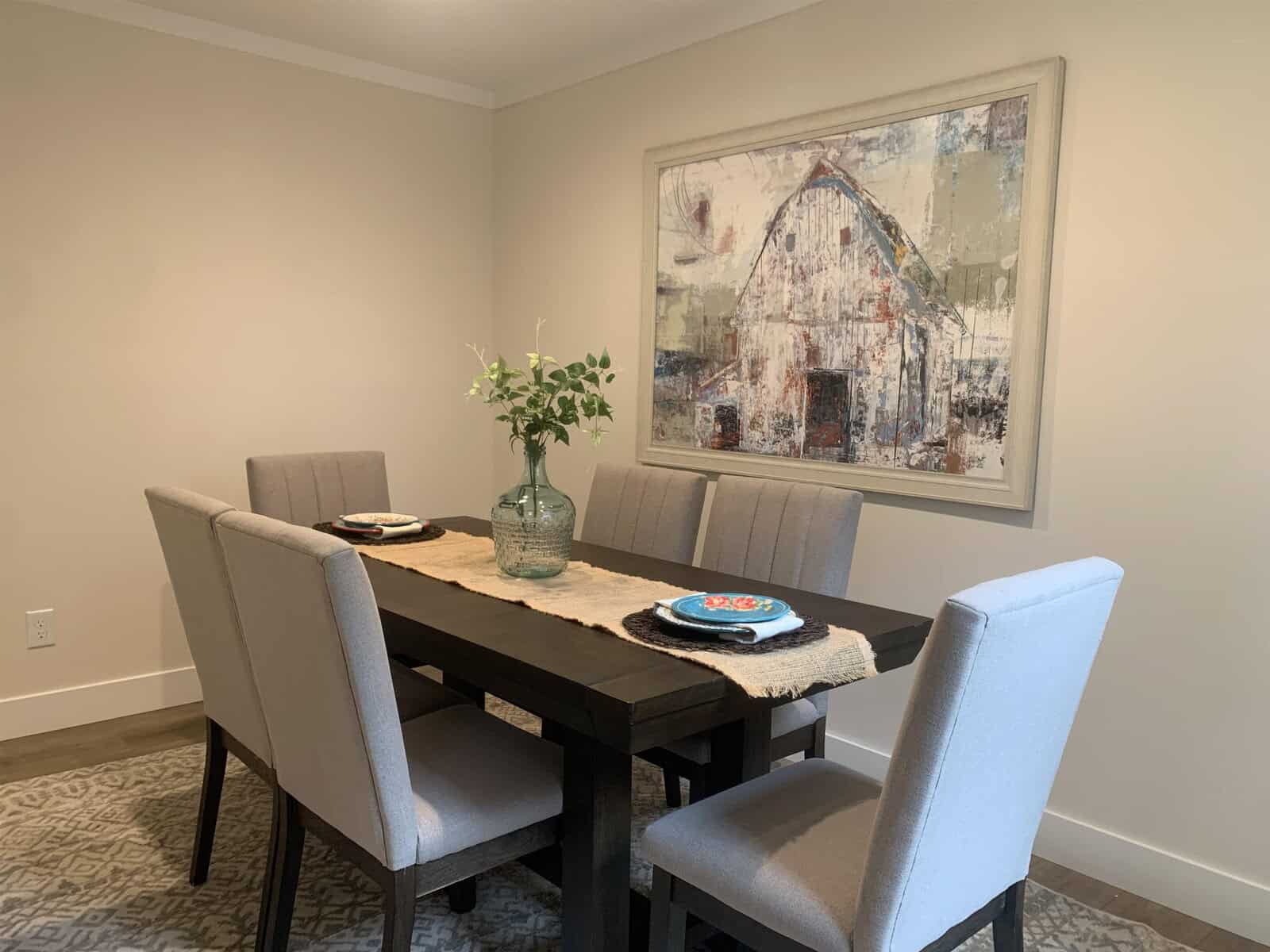 A modern dining area featuring a wooden table with four upholstered chairs, a centerpiece with green leaves, place settings for two, and a large abstract painting on the wall.