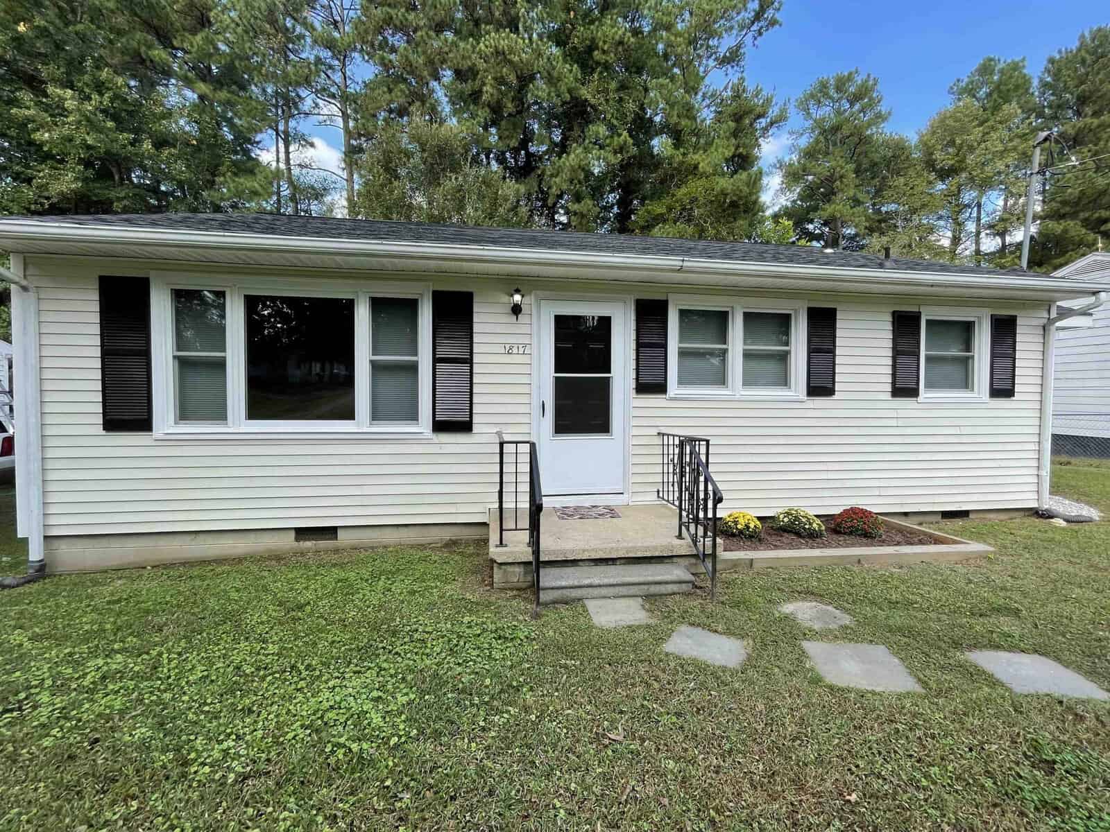 A single-story white house located at 1817 Willow Lane, Henderson NC, with black shutters, a gray shingled roof, and a small front porch with metal railings. Thereโs