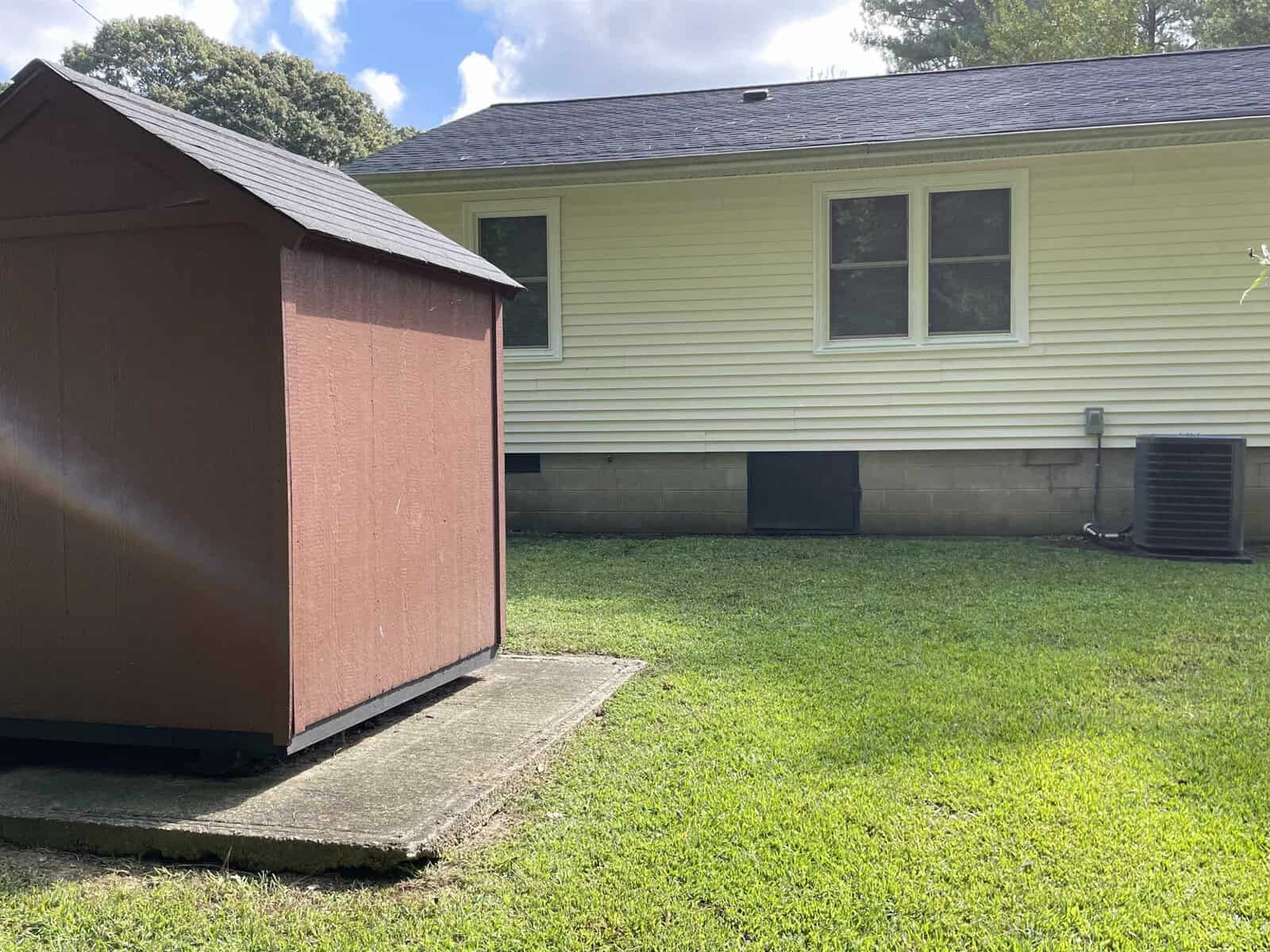 A small, brown shed beside a house with beige siding under a clear sky with visible air conditioning units and a well-maintained lawn.