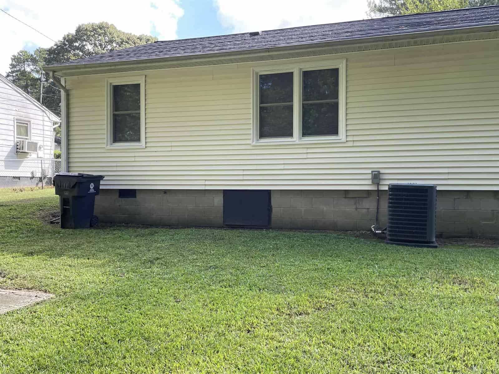 A single-story house with beige siding and a black foundation, featuring two windows, with a black trash bin and an air conditioning unit on the side.