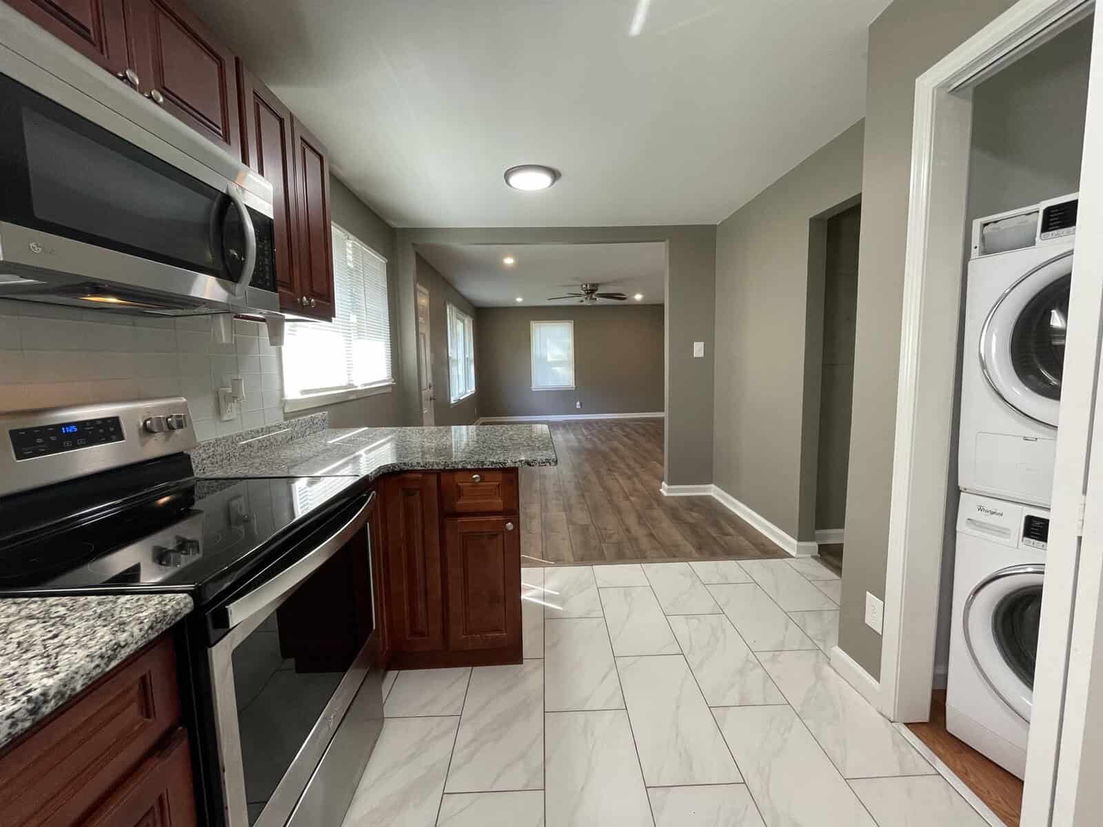 A modern kitchen with stainless steel appliances and granite countertops extends into a living area with hardwood floors, adjacent to a laundry area with a washer and dryer.