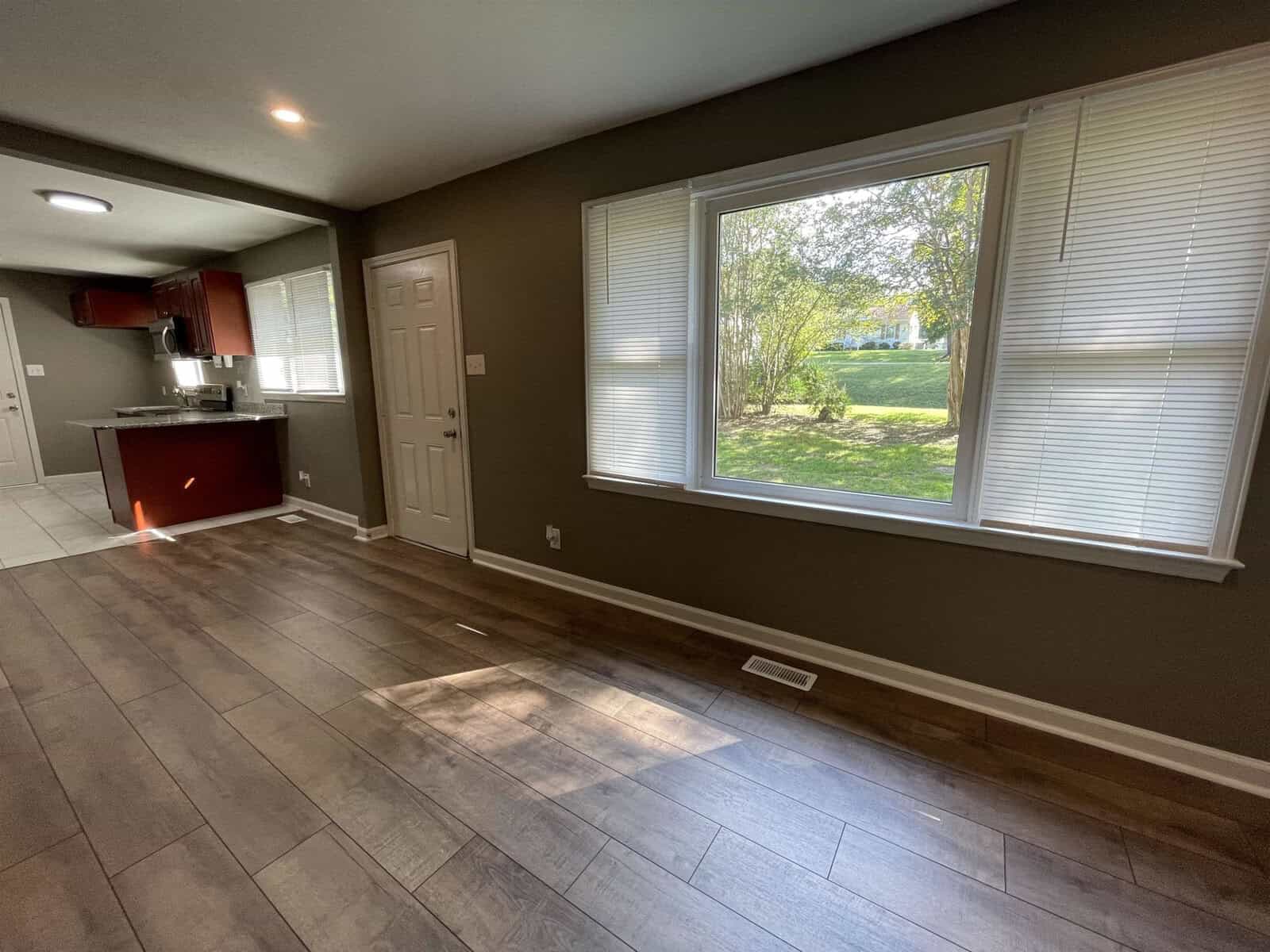 An empty room with wooden flooring, a brown wall, two large windows with blinds partially open, and a white door. a kitchen area is visible to the left with a countertop.