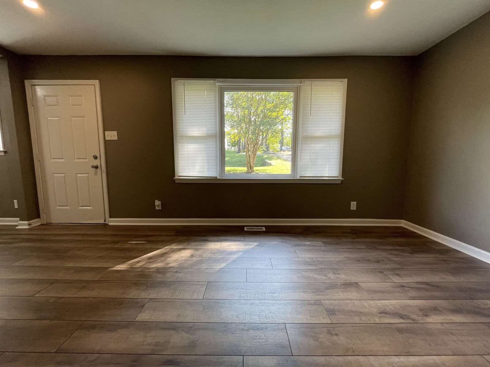 An empty room with dark wood flooring, a closed white door on the left, and a large window with blinds looking out onto a tree.