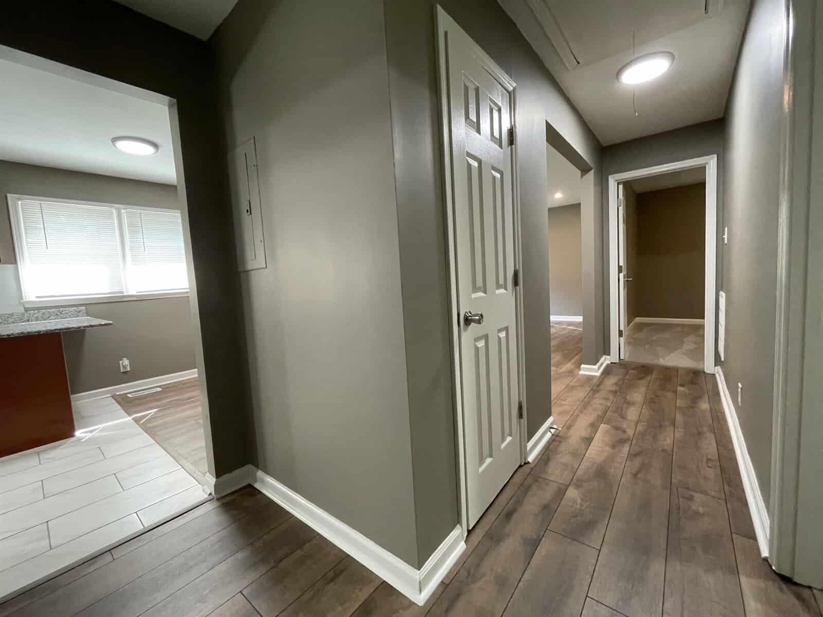 A view of a modern interior hallway featuring wooden floors, white doors, and neutral-colored walls, with a kitchen area visible to the left and another room accessible straight ahead.
