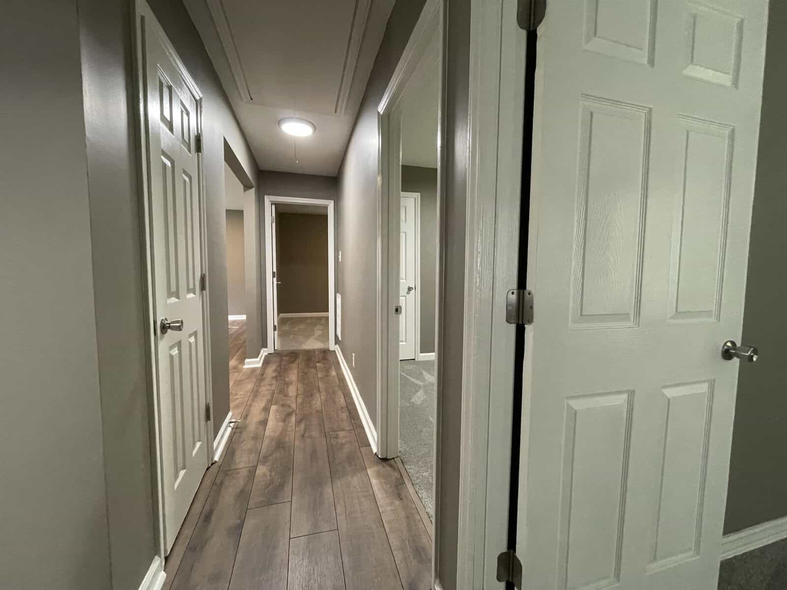 A residential hallway with multiple closed doors and wood-like flooring, illuminated by overhead lights.