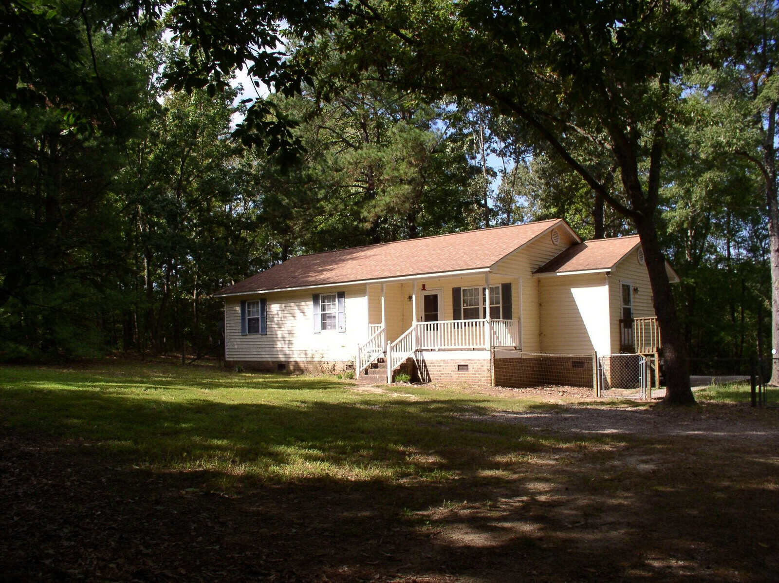 A single-story, white residential house with a gable roof, a front porch with stairs, and surrounded by trees and a lawn.