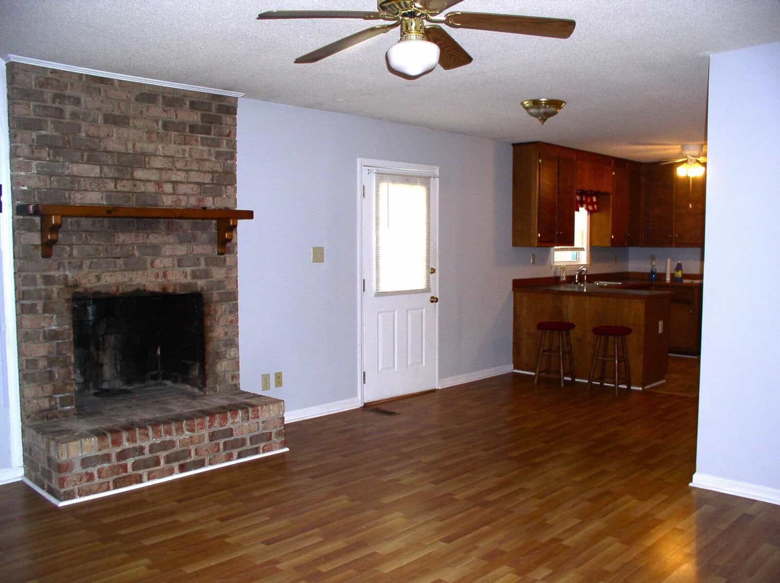 An empty living room with hardwood floors, a brick fireplace, and an adjoining kitchen area visible in the background.