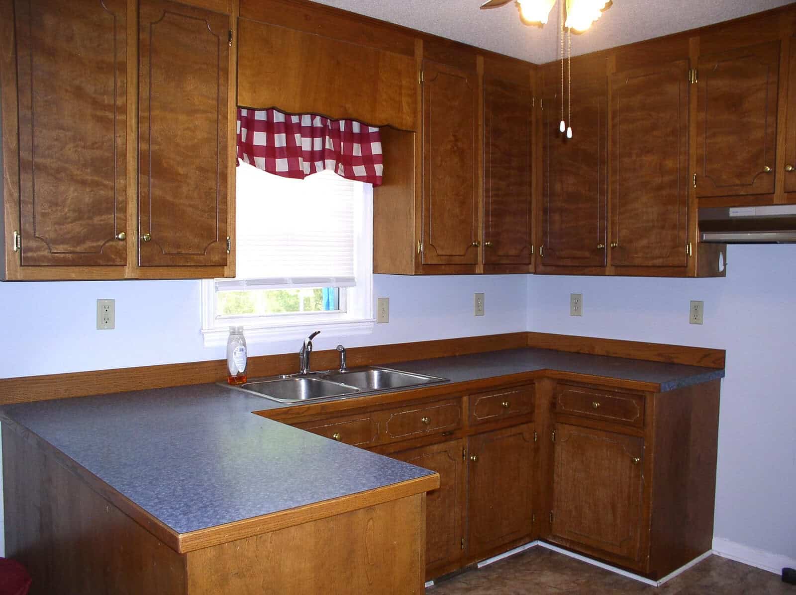 A clean, empty kitchen featuring wooden cabinets, laminate countertops, a double sink, and a window with a red and white checkered valance.