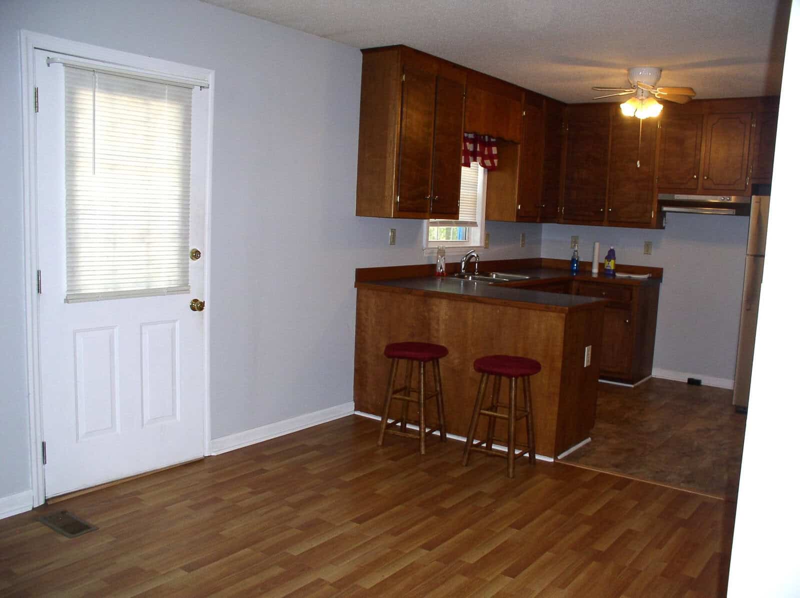 A tidy kitchen with wooden cabinets and flooring, featuring a breakfast bar with two red stools, and a door with a glass panel on the left.