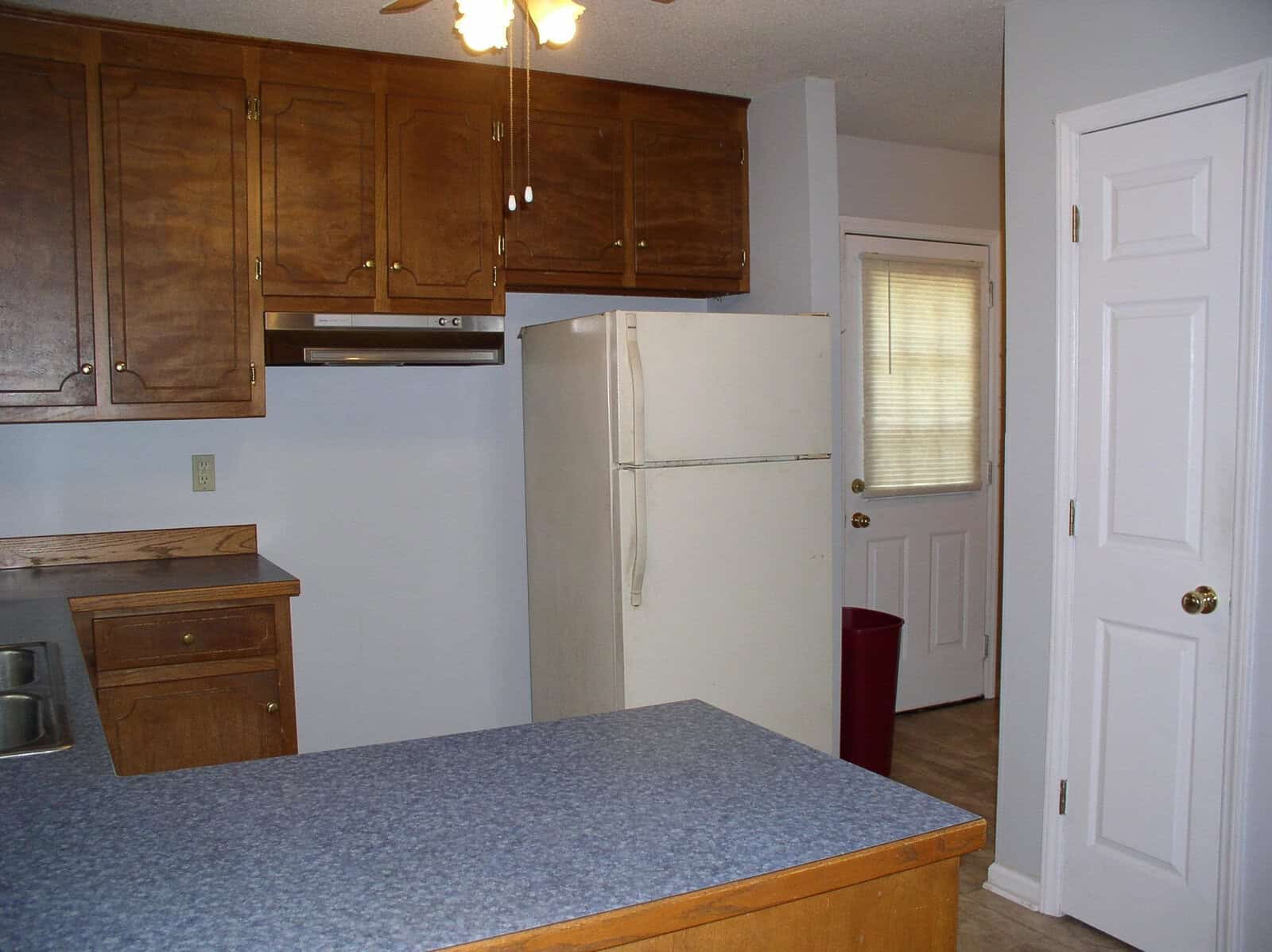 A modest kitchen interior with wooden cabinets, blue countertops, a white refrigerator, and a closed door on the right.