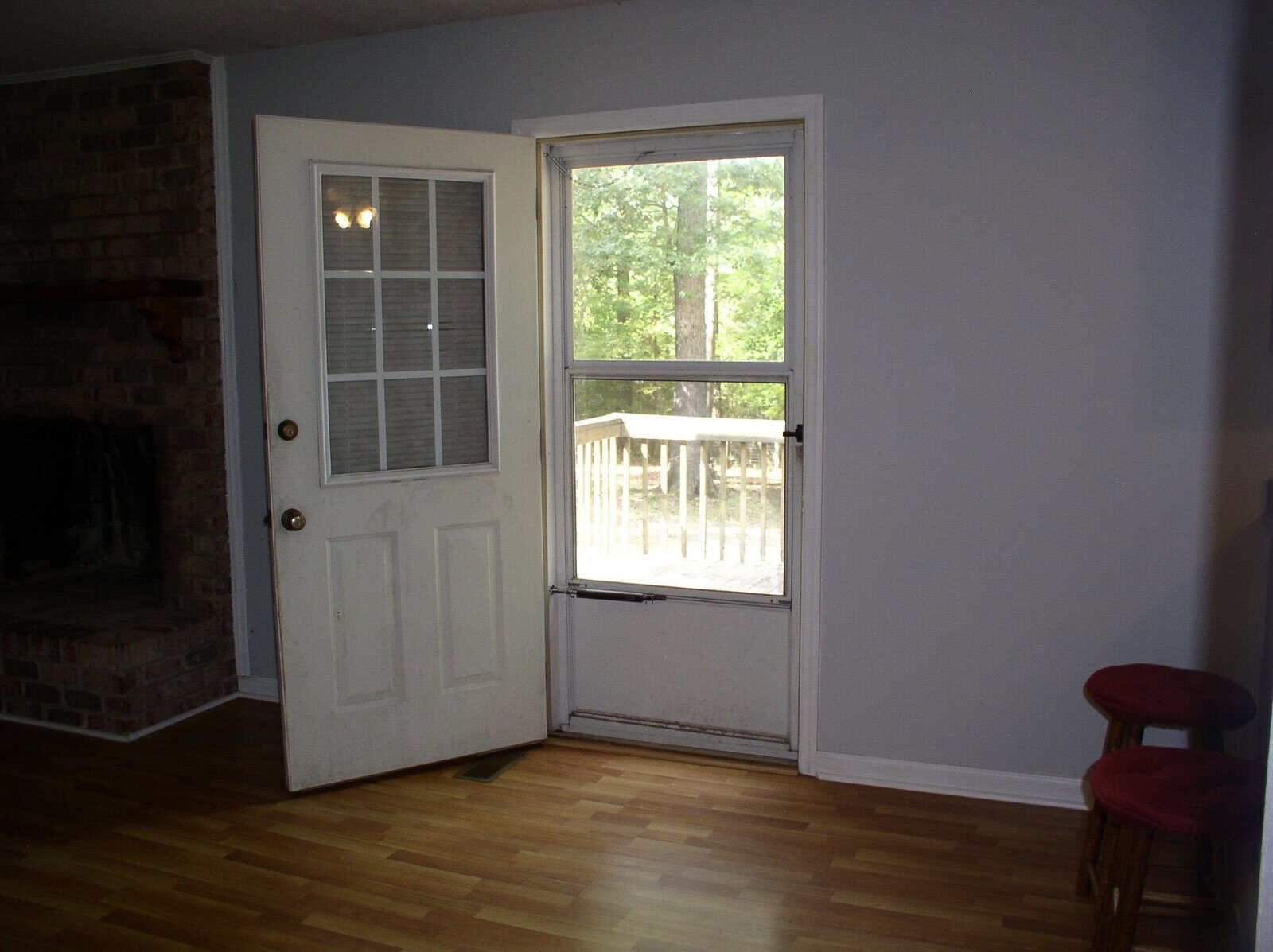 An empty room with wooden flooring featuring a white door to the left, a fireplace on the left wall, a large window with a view of trees, and a solitary red stool.