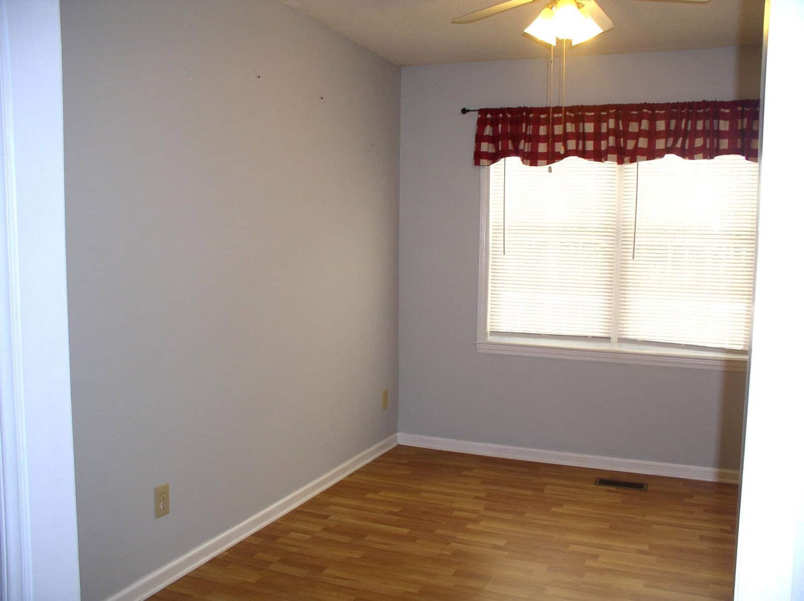 An empty room with beige walls, a window with a red valance, and hardwood flooring.
