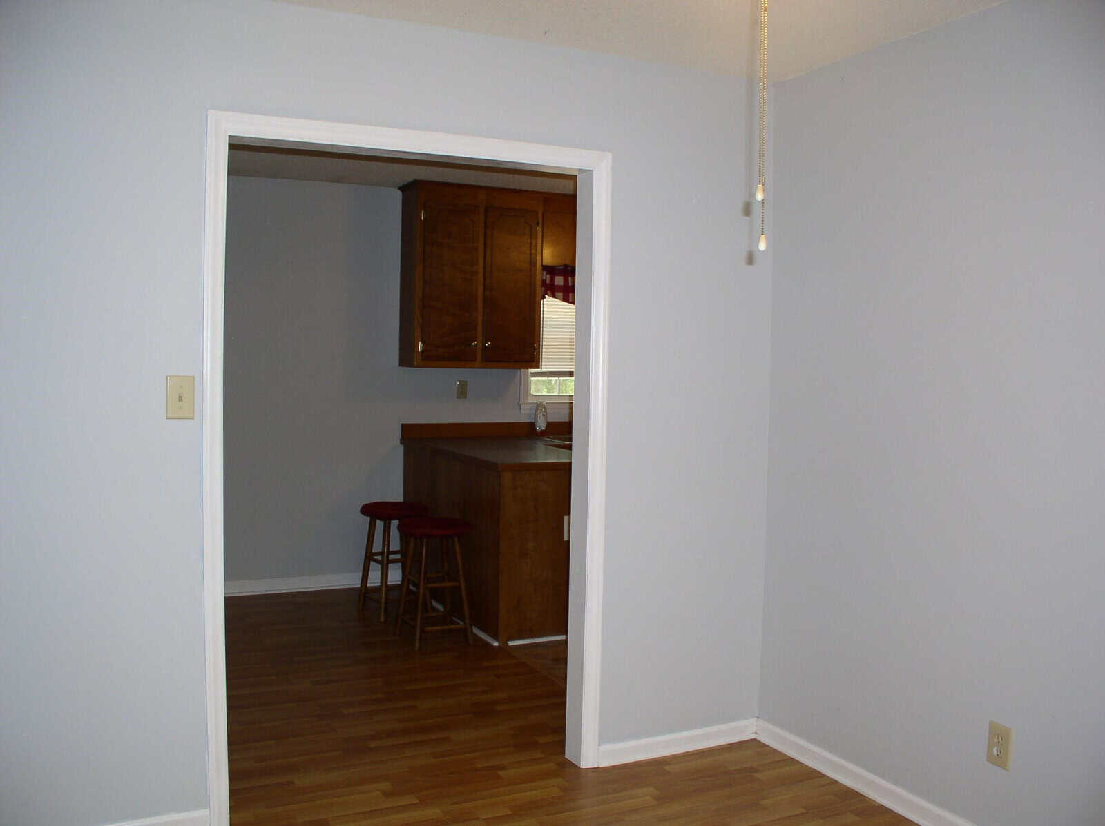 An empty room with gray walls and hardwood flooring leading to a kitchen with wooden cabinets and a red stool.