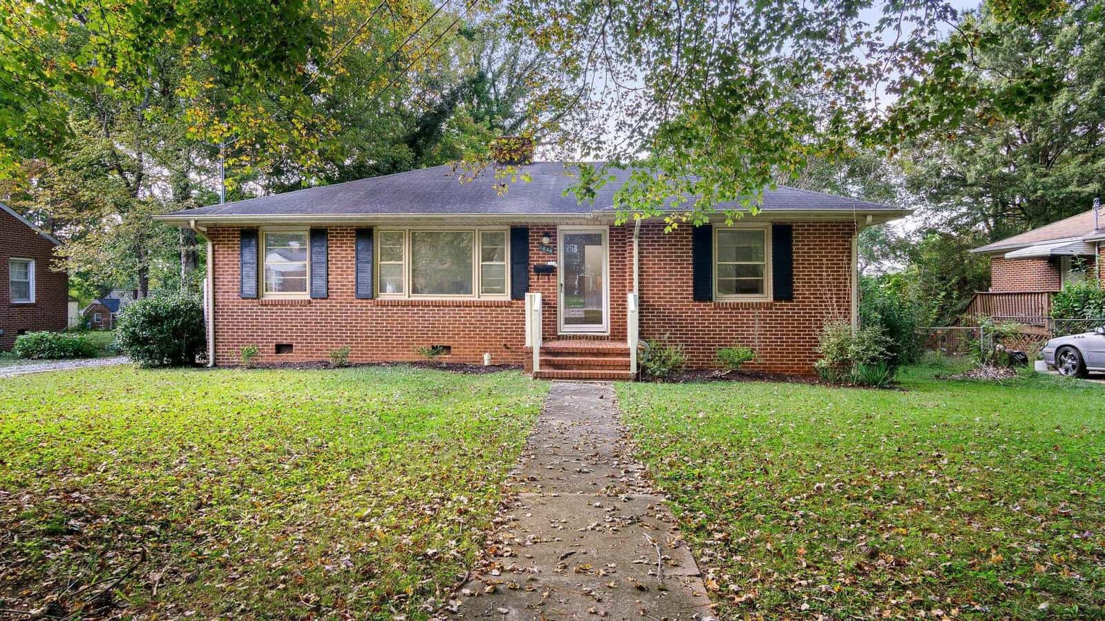 A quaint brick single-story house with a pitched roof and a front lawn, featuring a concrete pathway leading to the entrance.