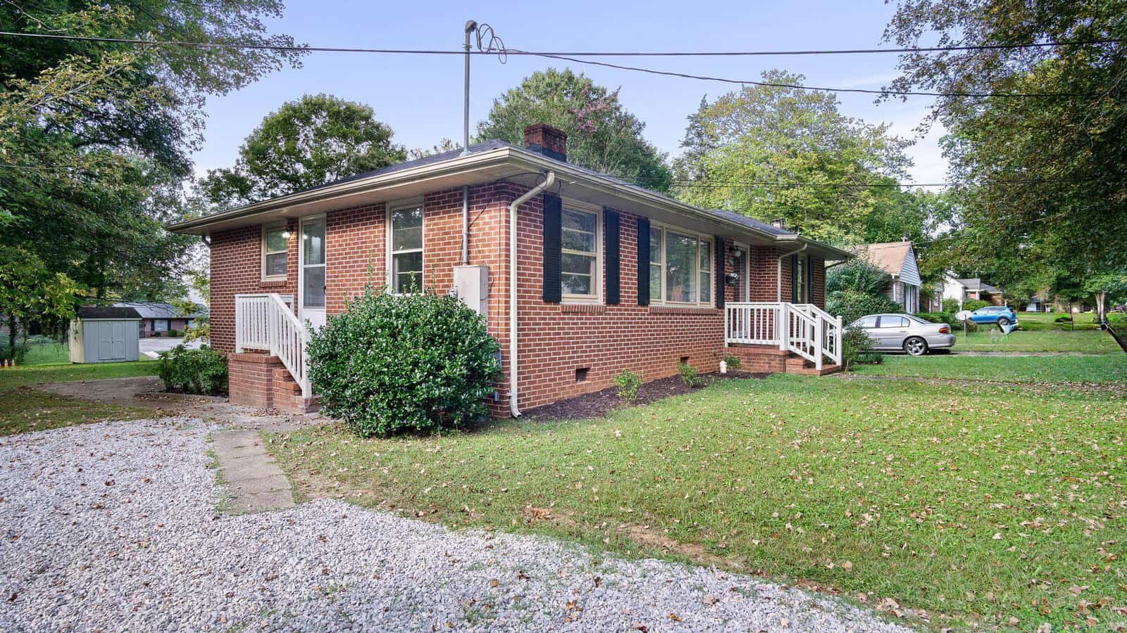 A single-story brick house with a white porch, surrounded by a lawn with a gravel driveway to the side.