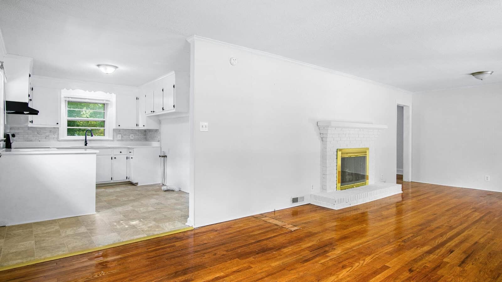 An empty, well-lit room with hardwood flooring featuring an adjoining kitchen with white cabinets and a fireplace with a white mantel and yellow-tiled hearth.