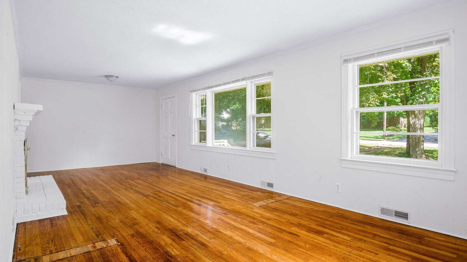 An empty room with polished hardwood floors, white walls, and three windows providing a view of the outdoor greenery.