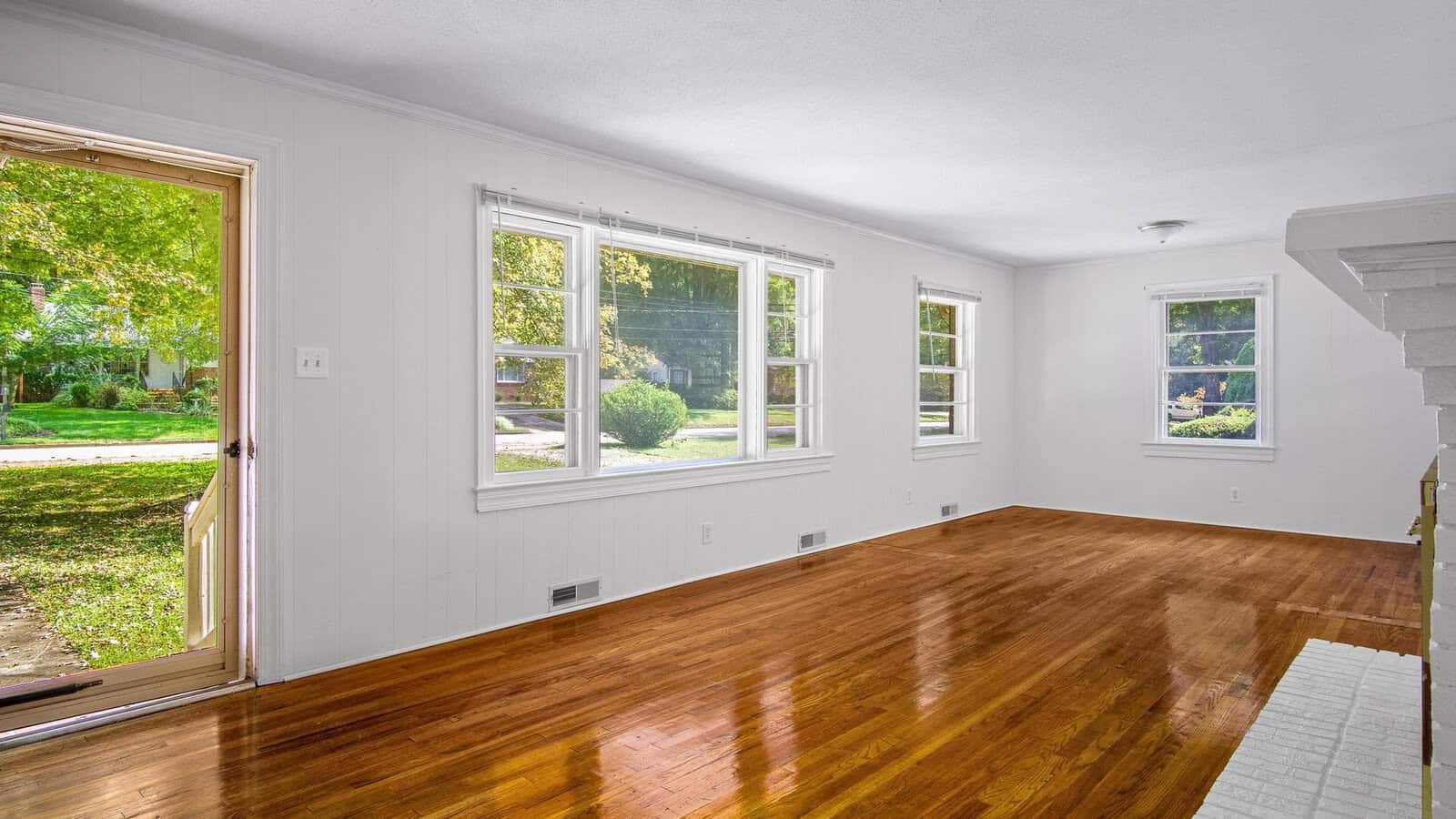 An empty room with polished hardwood floors, white walls, and three windows providing ample natural light.