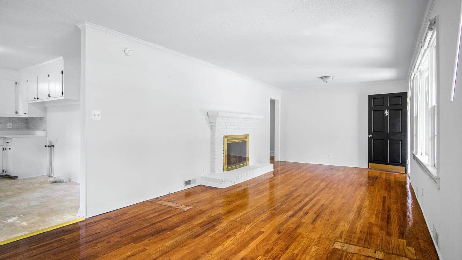 An empty room with hardwood floors, white walls, a fireplace, and a view into a kitchen with white cabinetry.