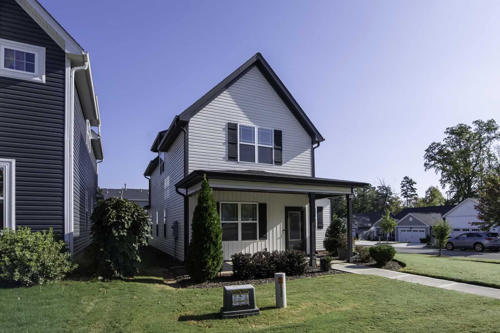 A two-story, light gray house with a dark roof is shown, surrounded by a well-maintained lawn and shrubs. A small pathway leads to the front door, and a neighboring house is partially visible on the left. The sky is clear and sunny.