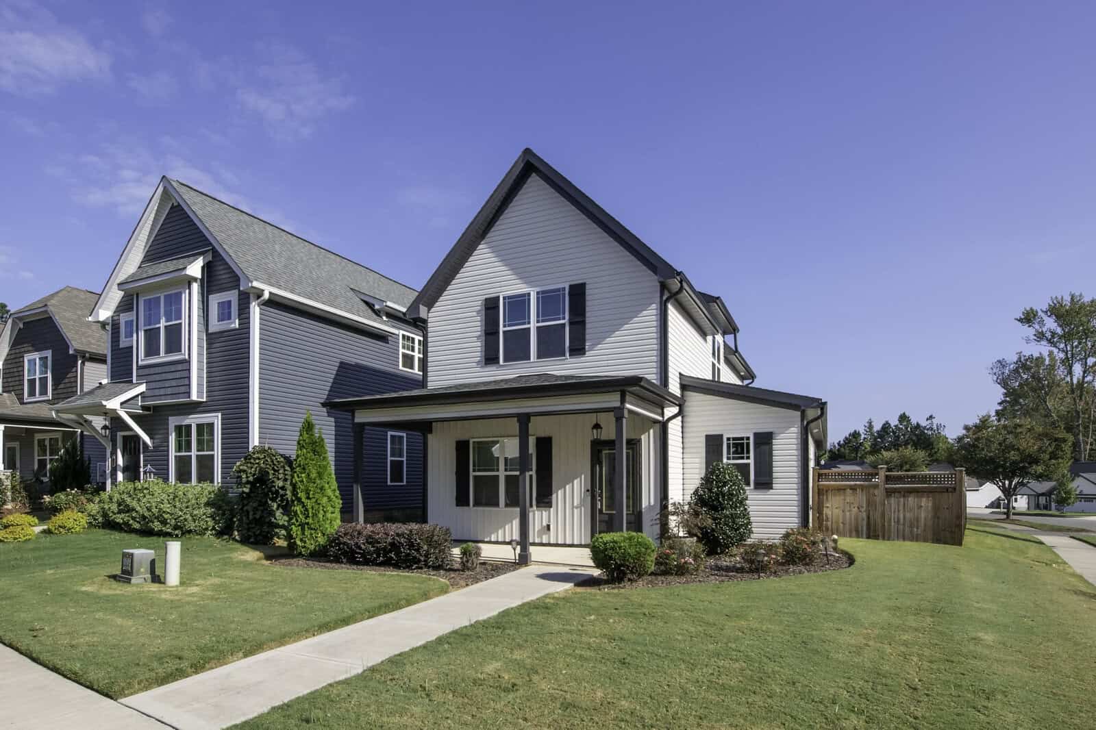 A modern suburban house with a gray exterior and a small front porch sits on a well-maintained lawn. It is part of a row of similar homes under a clear blue sky. A sidewalk runs along the front yard, and a wooden fence marks the backyard area.