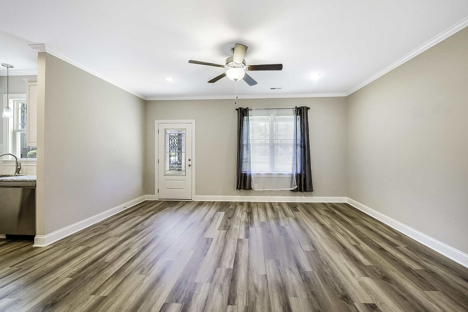 A spacious, empty room with light wood flooring, beige walls, and a ceiling fan. A window with sheer curtains and a glass patio door are on one wall, and part of a kitchen area is visible to the left.
