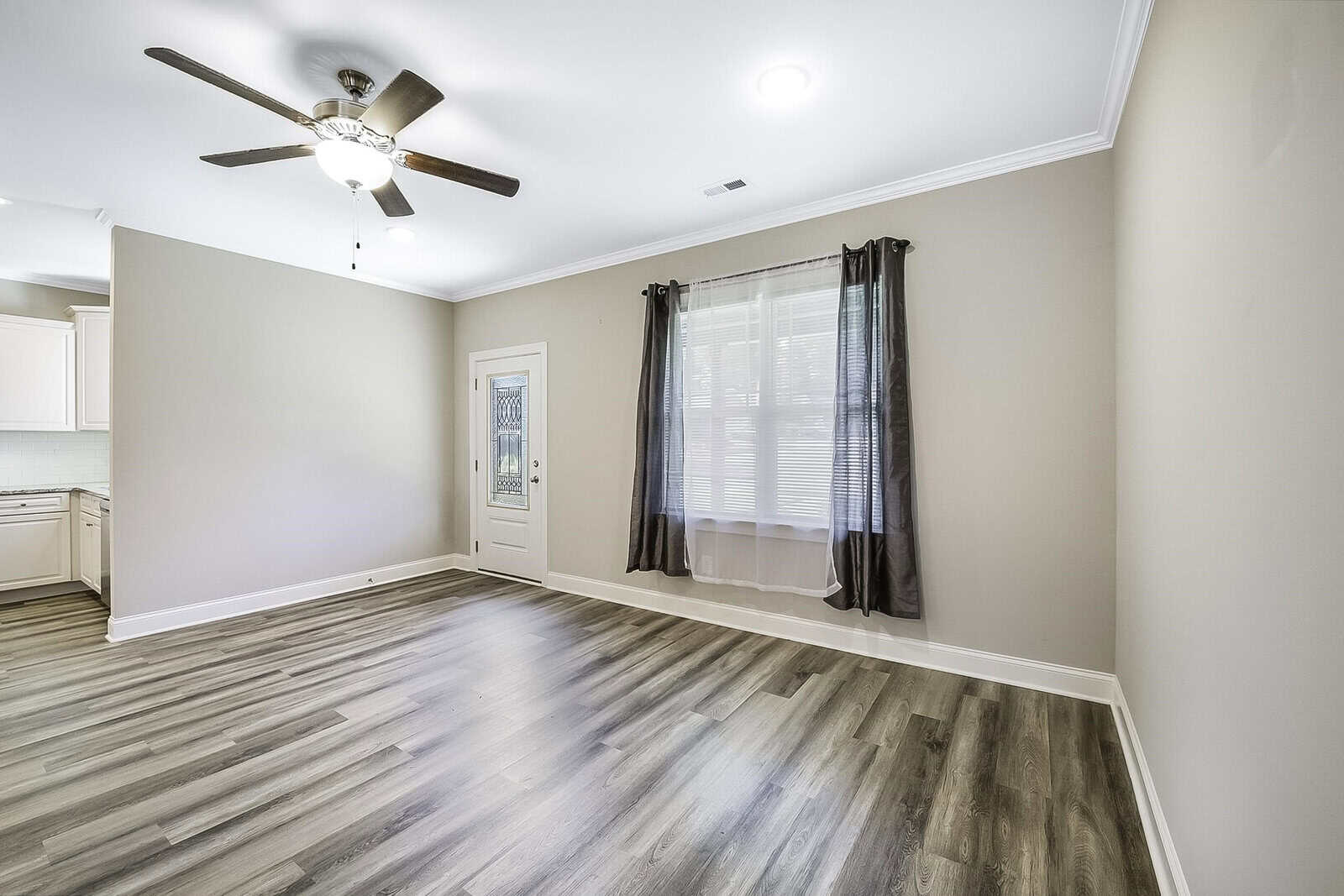 A spacious, empty room with light gray hardwood flooring, beige walls, and a ceiling fan with a light. There is a large window with dark curtains and a door leading to another room. The room is well lit.