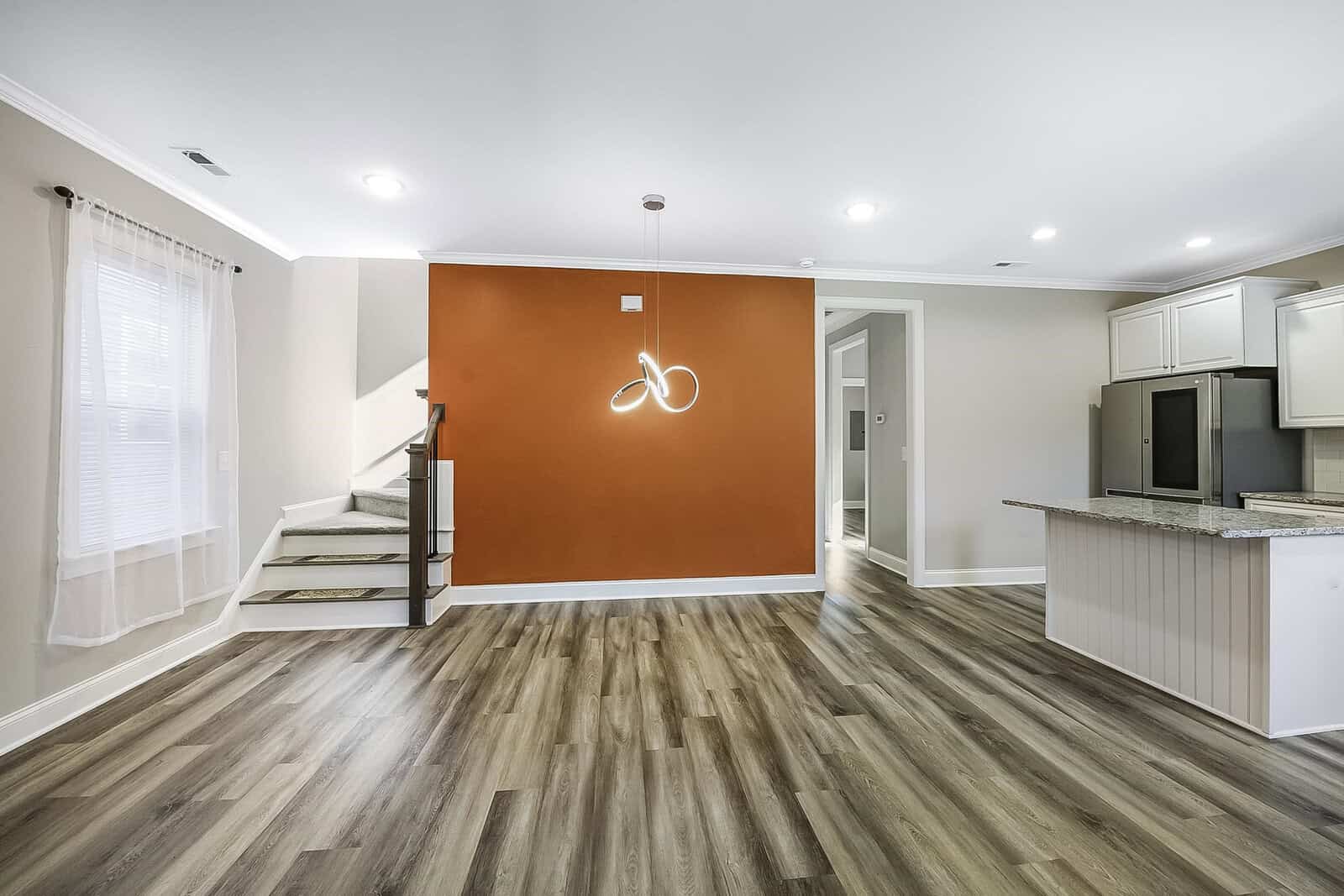 A modern, open living space with a warm orange accent wall and a contemporary light fixture. Features wood flooring, white cabinetry, and a kitchen area with stainless steel appliances. A staircase leads to the upper level.