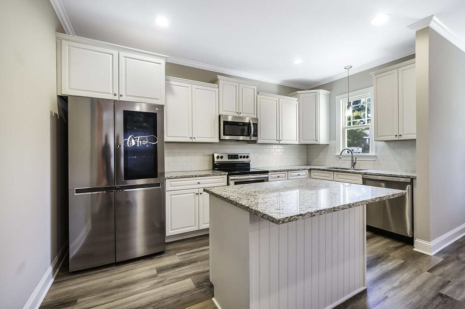 Modern kitchen with stainless steel appliances, including a fridge and microwave. Light-colored cabinets and a granite-topped island stand out against a neutral backsplash. Hardwood floors add warmth to the space. A window brings in natural light.