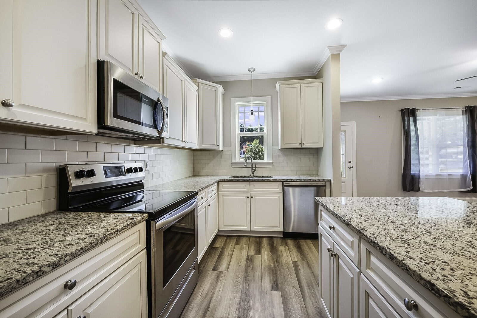 Modern kitchen with white cabinets, stainless steel appliances, and granite countertops. The space includes a stove, microwave, and dishwasher. Hardwood floors and a window above the sink add to the bright, clean look. Curtains partially cover a window.