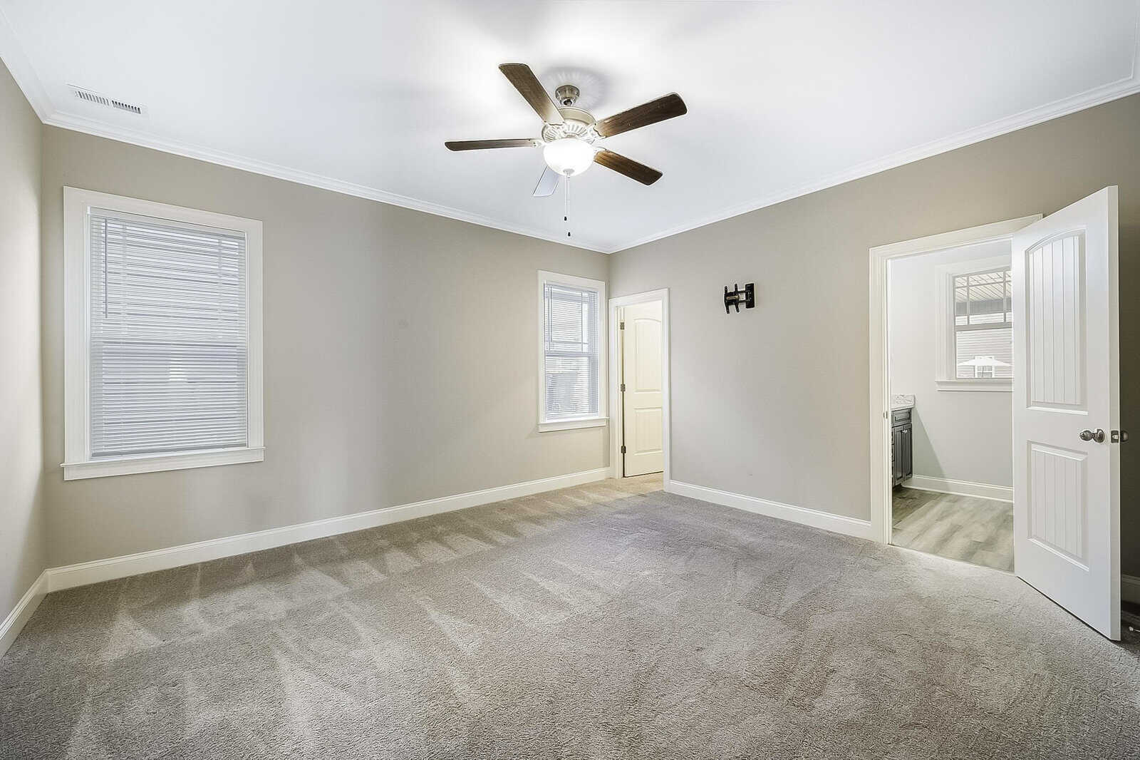 A spacious, empty room with gray carpet, beige walls, and a white ceiling. There is a ceiling fan with a light in the center. Two windows with blinds are on one wall, and an open door leads to another room with a visible washer and dryer.