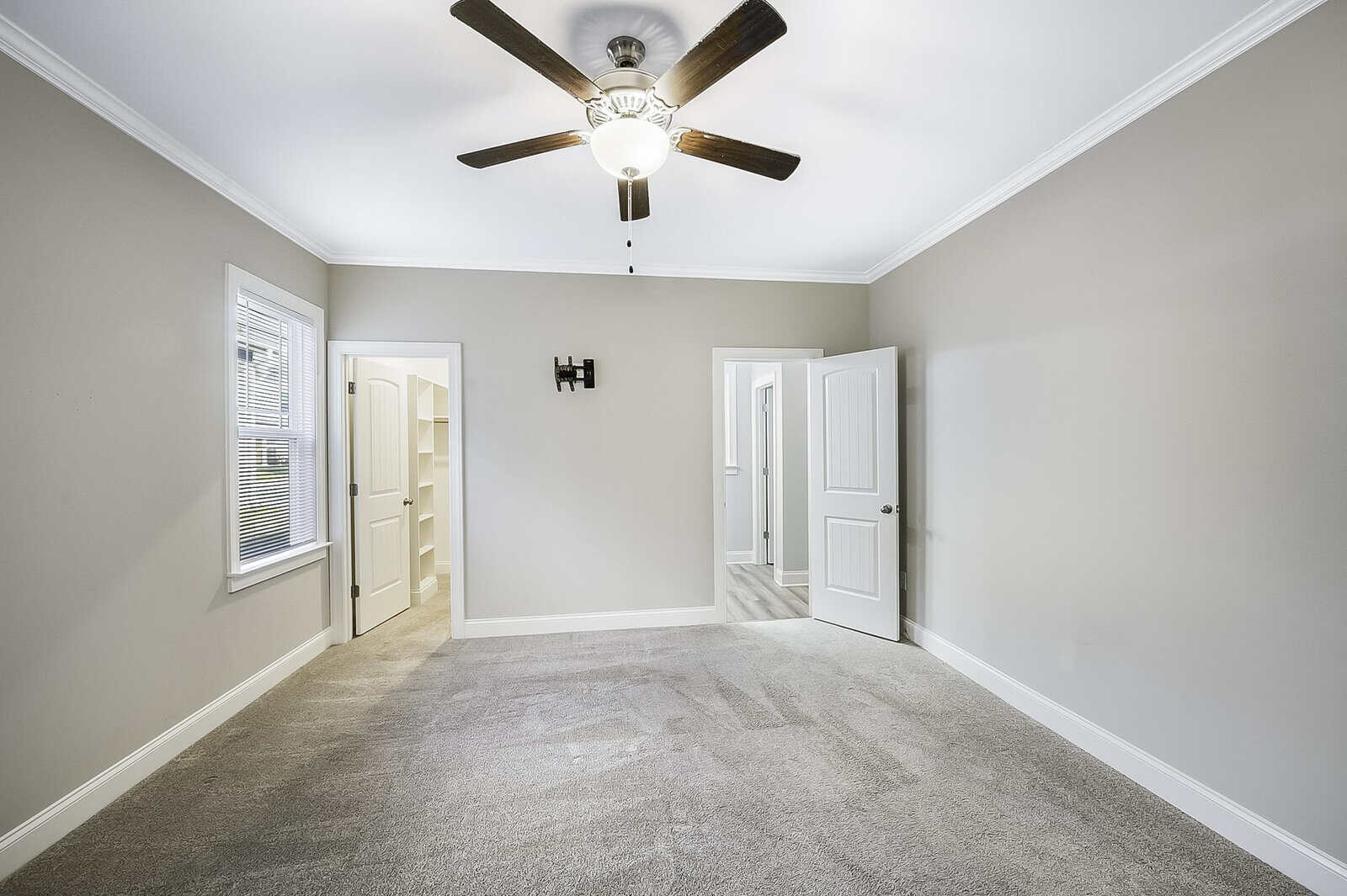 A bright, empty room with beige walls and carpeted floor. A ceiling fan with light is centered above. The room features a window on the left, a walk-in closet with shelves, and an open door leading to another room.