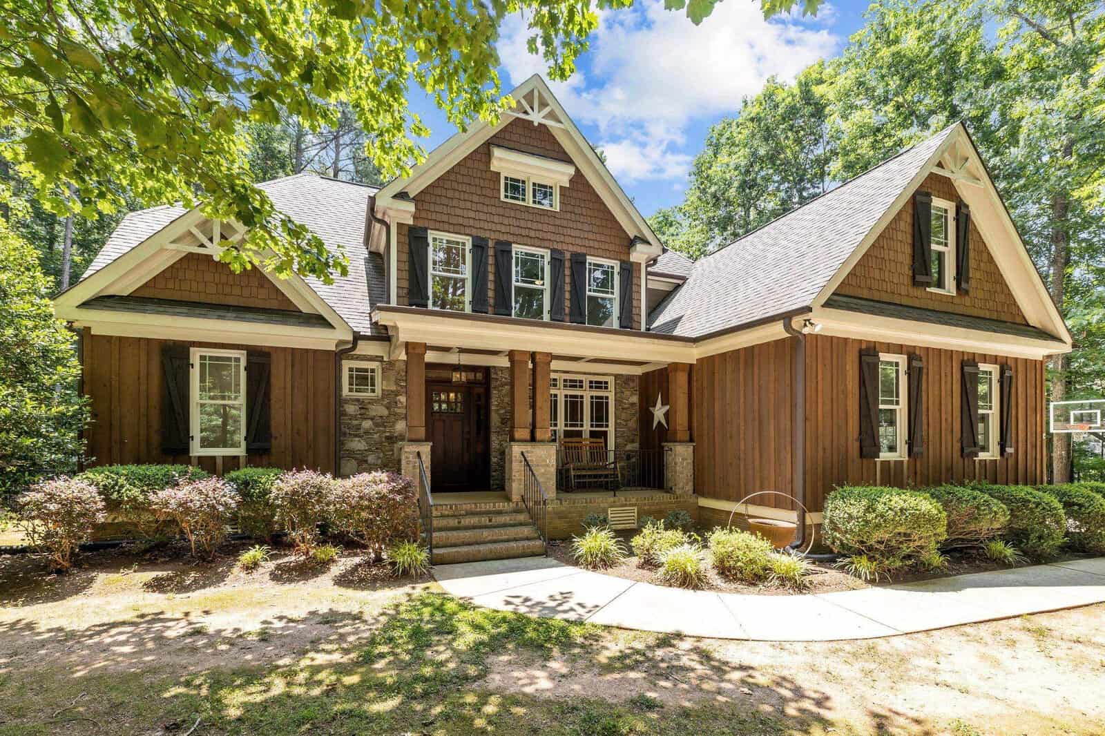 A charming two-story house at 1194 Old Still Way, Wake Forest, boasts brown wooden siding, white trim, and a stone facade. It features dormer windows, a gable roof, and a well-maintained garden with hedges and trees under a clear blue sky.