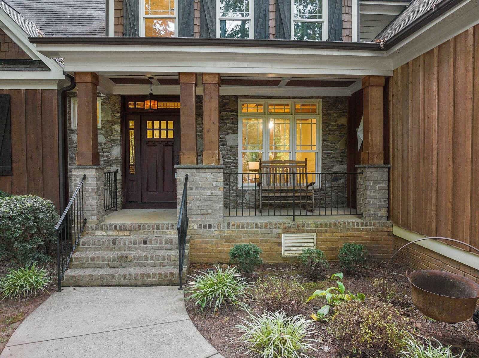 A welcoming front porch of a house with a dark wooden door, large windows, and brick steps. There's a wooden bench on the porch, surrounded by lush plants and shrubs. The exterior is a mix of wood and stone.