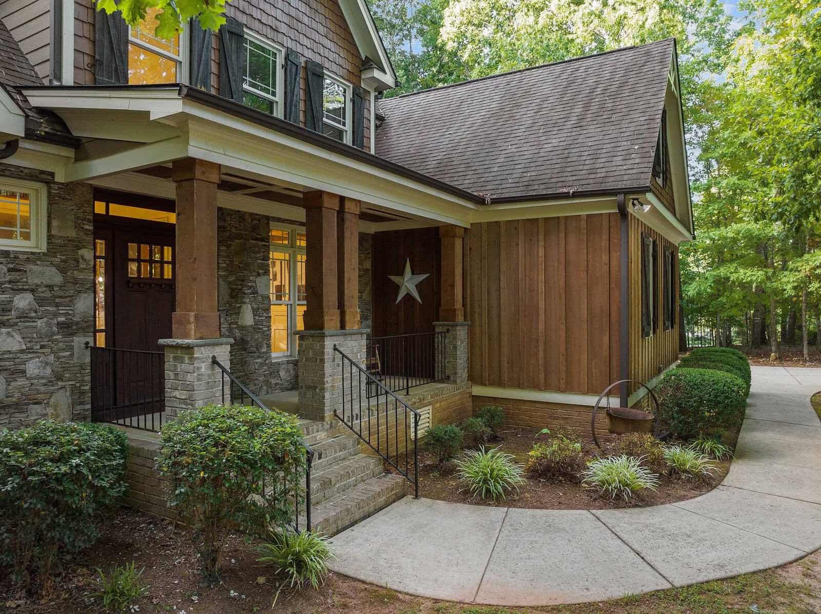 Front view of a rustic house with stone and wood siding. It features a covered porch with stairs and metal railings, a decorative star on the wooden wall, and surrounding greenery. A curved pathway leads to the entrance.