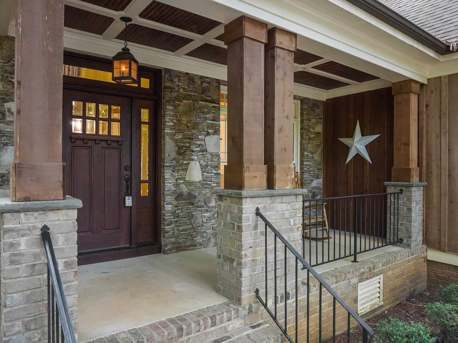 A rustic front porch with brick and stone walls, featuring a wooden door with glass panels, a hanging lantern, and a large star decoration on the wall. The porch has metal railings and wooden pillars supporting the overhang.