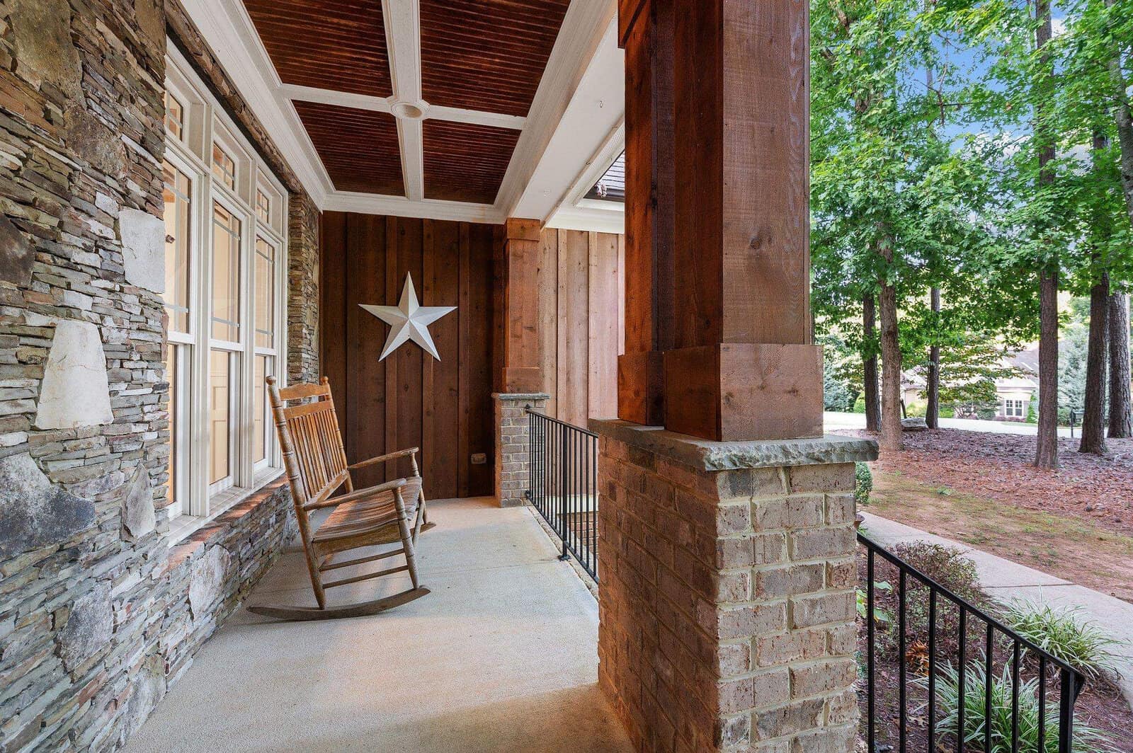 A rustic porch with wooden and stone elements features a wooden rocking chair and a star decoration on the wall. The porch overlooks a landscaped garden with trees.