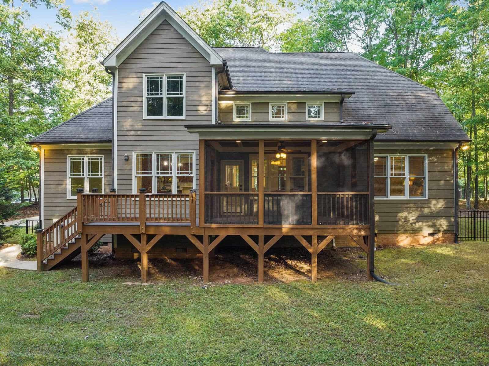 A two-story house with gray siding and a dark roof, featuring a screened-in porch and a wooden deck. Surrounded by trees, the yard is grassy and spacious, offering a serene natural setting.