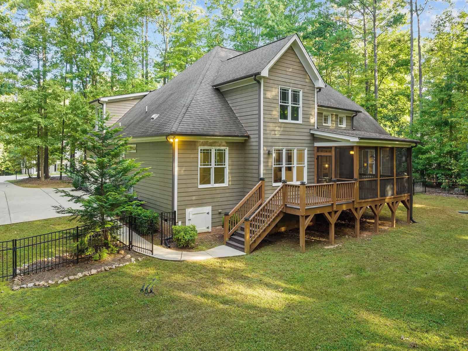 A two-story house with a gray exterior, surrounded by a lush green lawn and trees. It features a screened porch on a wooden deck and a fenced backyard. The driveway is partially visible on the left.