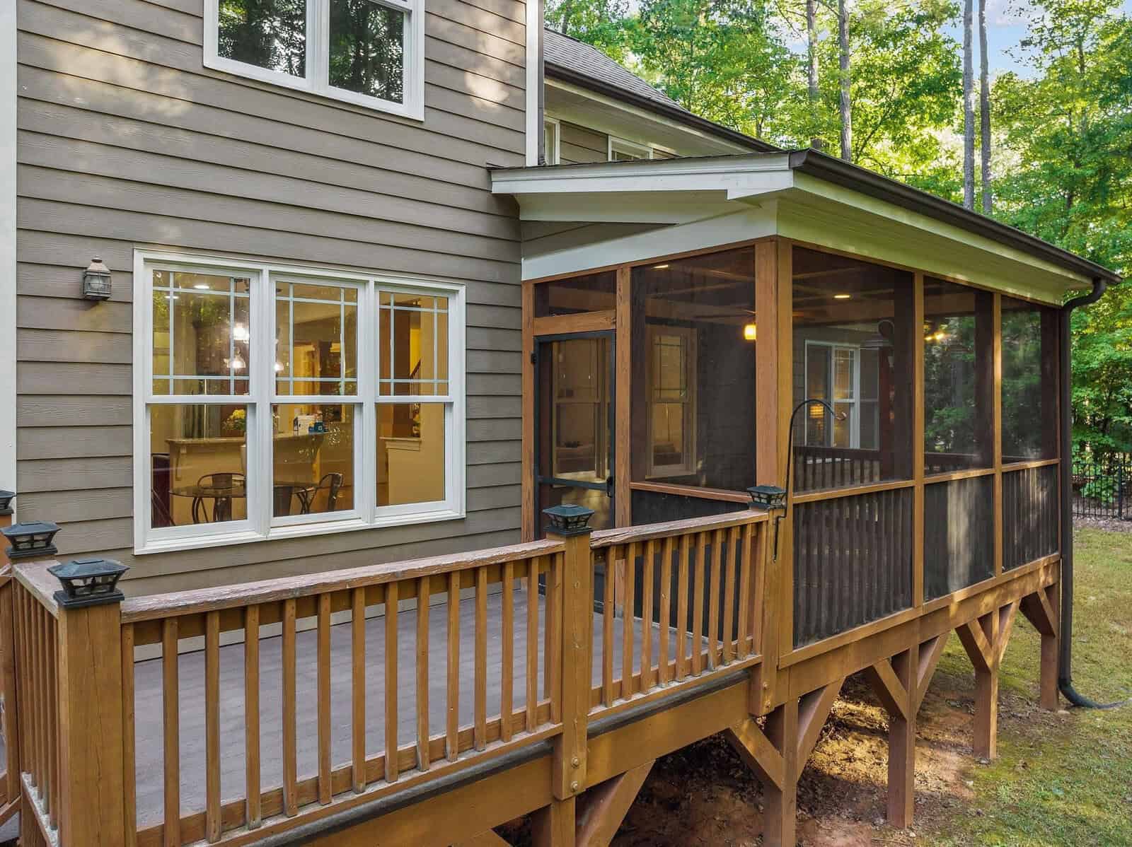 A house exterior with a brown wooden deck and a screened-in porch. Large windows reveal the home's interior, and the surrounding area is wooded with green trees.