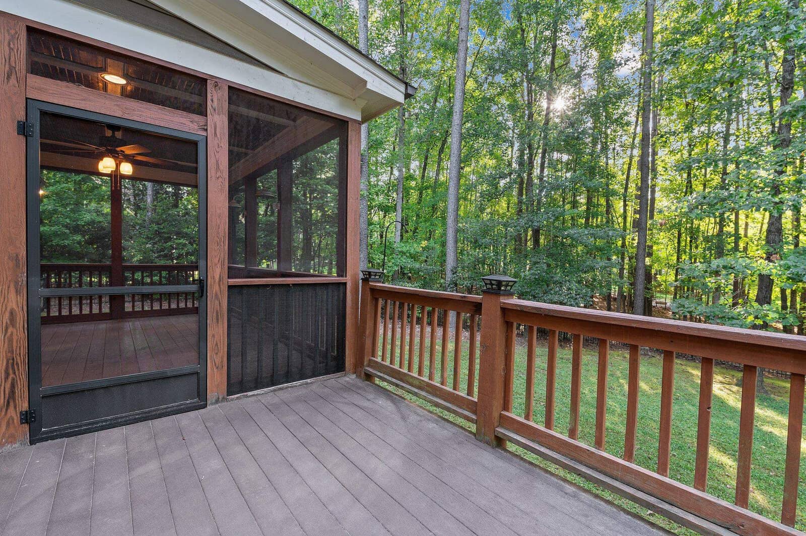 A wooden deck with a screened-in porch featuring a ceiling fan. The porch overlooks a lush green forest, letting in filtered sunlight through the trees. The deck has a railing and opens up to a grassy yard.