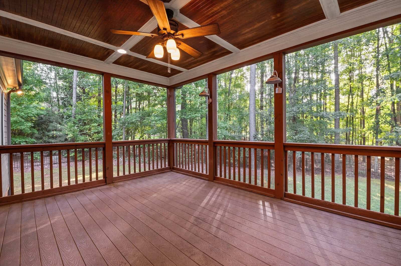 A spacious wooden deck with a ceiling fan and screened walls overlooking a lush forest. The tall trees and green foliage surround the serene outdoor space, providing a peaceful and natural setting.