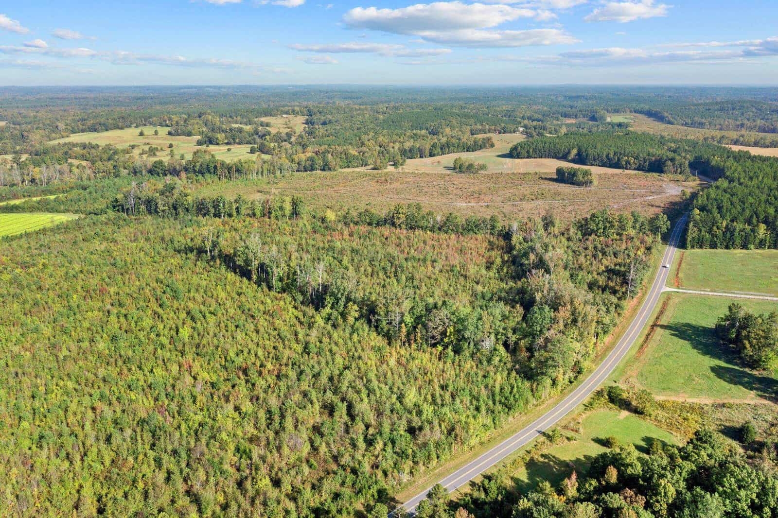 Aerial view of a rural landscape featuring dense green forests, open fields, and a winding road cutting through the terrain. The sky is partly cloudy, with patches of blue visible above the expansive countryside.
